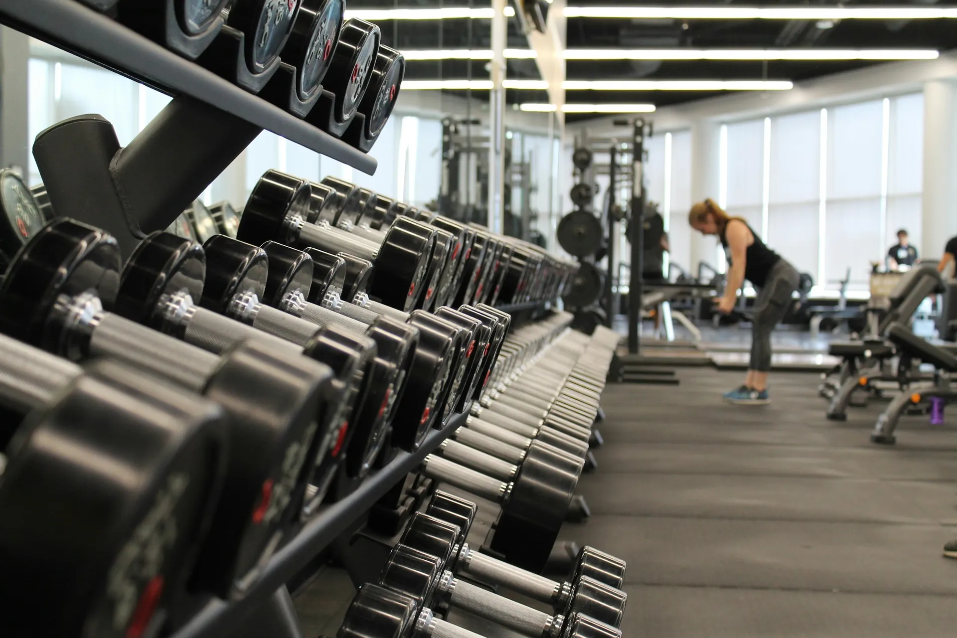 Salão de musculação com fileiras de halteres e alunos treinando ao fundo
